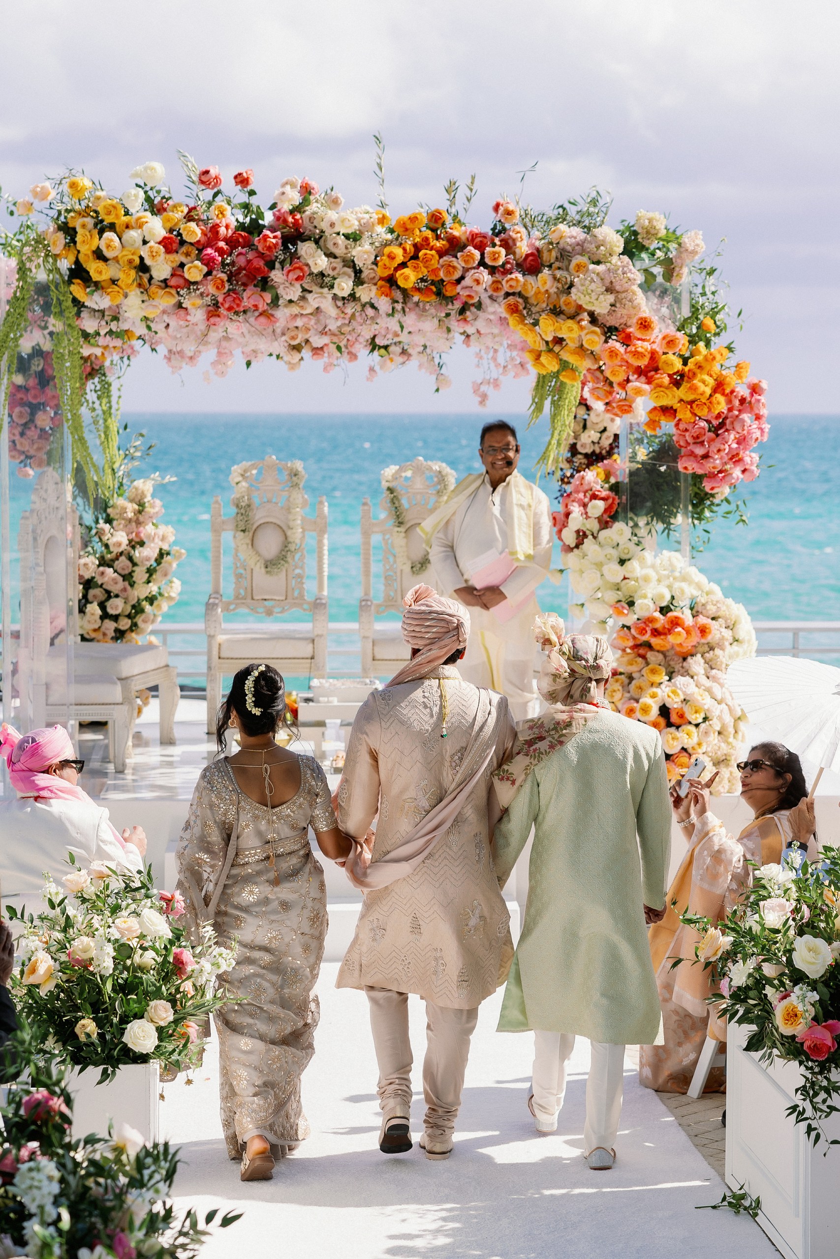 Groom being escorted down the aisle by his parents to a colorful mandaap in front of the ocean